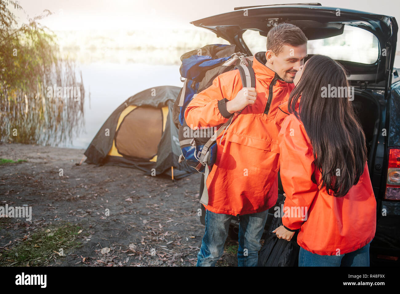 Schönes Bild von junges Paar küssen. Sie tragen orange Jacken. Menschen, die in der Nähe von einander. Er hat Rucksack. Paar ist bei geöffneten Kofferraum und Zelt. Stockfoto