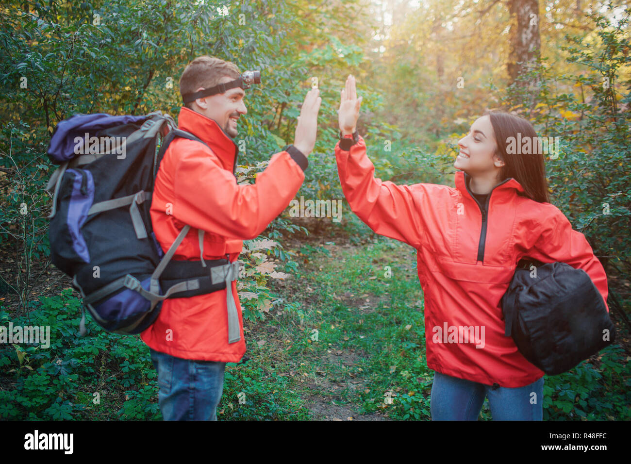 Junge Mann und die Frau in der orangefarbenen Jacken fünf Zeichen zueinander. Er hat Rucksack. Sie hält Schlafsack. Sie lächeln. Menschen sind im Wald. Stockfoto