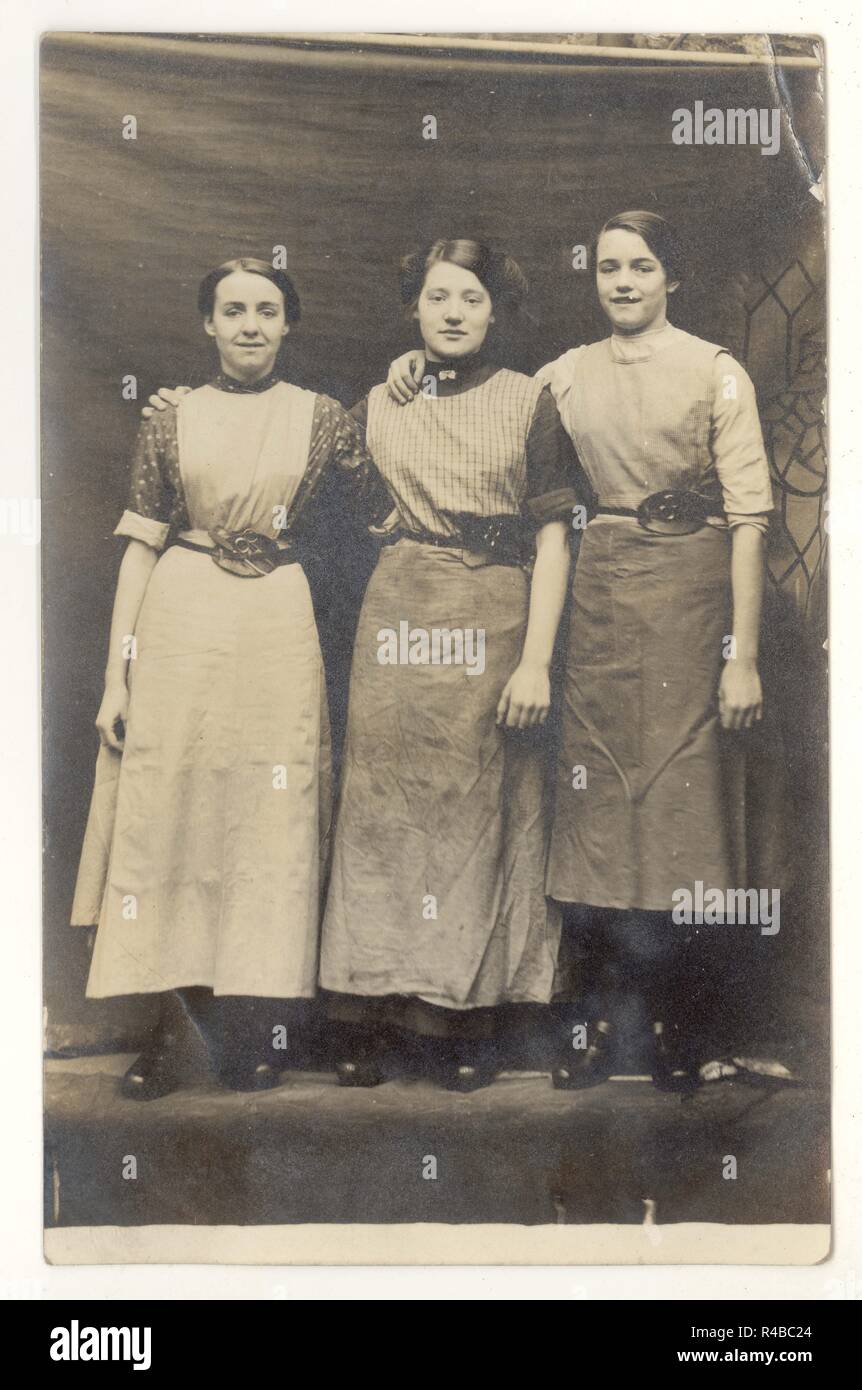 WW1 Studioporträt aus der Ära von 3 Mädchen, möglicherweise Arbeiter der Baumwollfabrik von Lancashire, die in ihren speziell entwickelten Gürteln Clogs und Schürzen mit einem hakenähnlichen Werkzeug und einer Schere trugen, um 1915,1918, Großbritannien Stockfoto