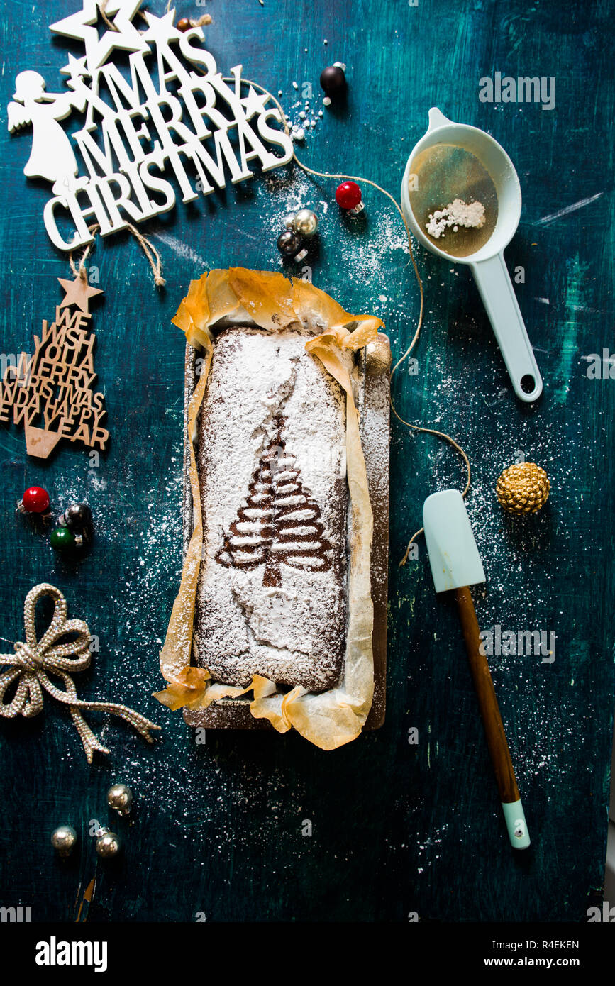Schokolade Banane Brot mit einem Weihnachtsbaum Dekor auf der Oberseite, Spatel, Sieb und Weihnachten Dekoration füllen Sie das Styling, flach über eine rustikale gr Stockfoto