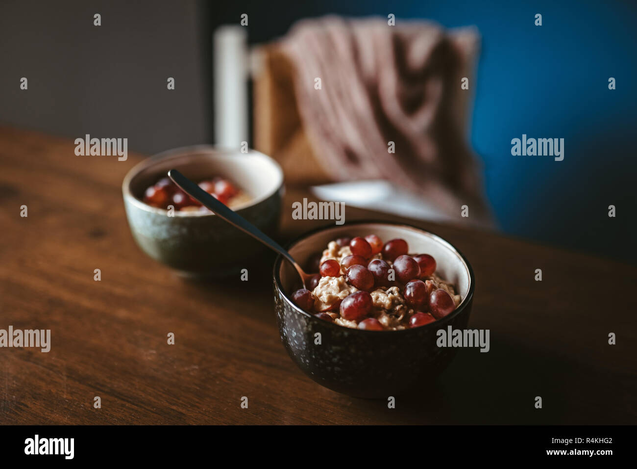 Zwei Schalen mit Porridge mit Trauben auf einen hölzernen Tisch mit Stuhl und Decke im Hintergrund Stockfoto