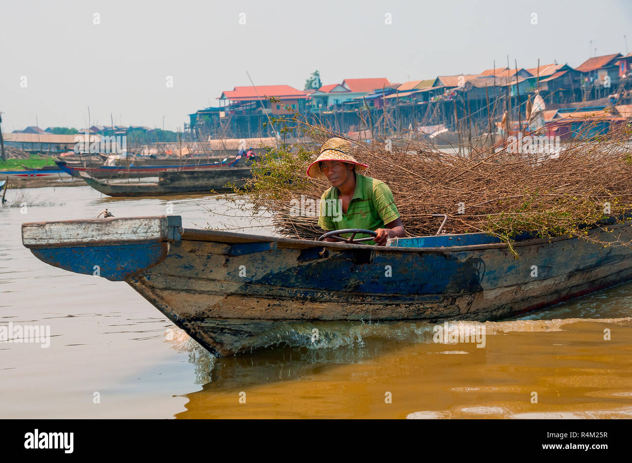 Mann, der traditionellen Holz- Motorboot vor der hölzernen Stange Häuser am Ufer einer Flussmündung anschließen an See Tonle Sap, Kambodscha gelegen Stockfoto
