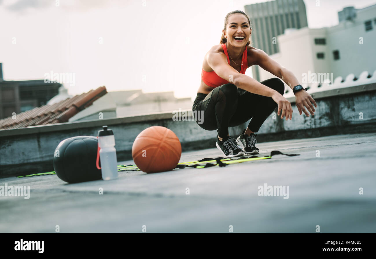 Fröhliche Frau in Fitnesskleidung beim Training auf der Terrasse. Fitness-Frau, die auf dem Dach sitzt, mit Medizinball und Basketball an ihrer Seite. Stockfoto