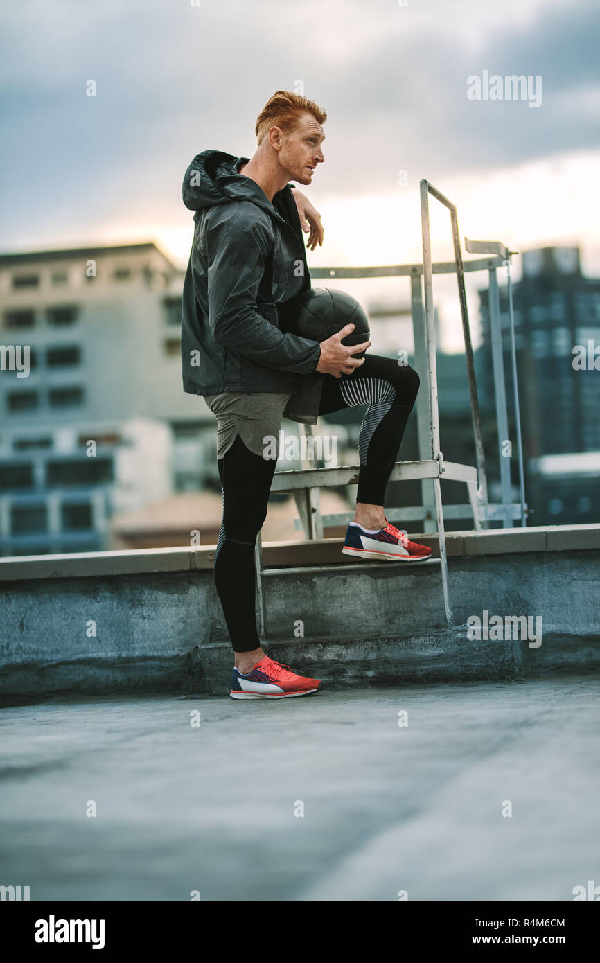 Seitenansicht eines Athleten in der Nähe von eine Dachterrasse mit Treppe und Wegsehen. Mann in Fitness Kleidung ständigen entspannt Halte einen Medizinball auf dem Dach Stockfoto