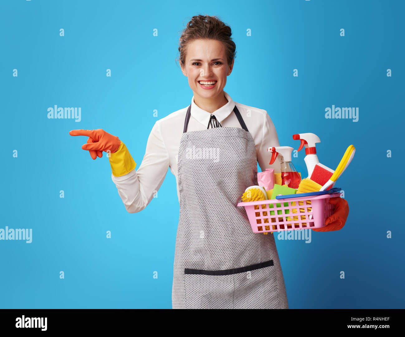 Lächelnde junge Putzfrau in Schürze mit einem Korb mit Reiniger und Bürsten an etwas vor blauem Hintergrund zeigen. Weibliche cleaner präsentiert Stockfoto