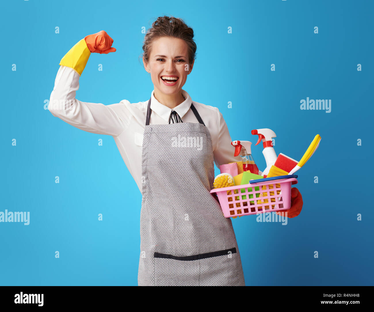 Glückliche junge Putzfrau in Schürze mit einem Korb mit Reiniger und Bürsten mit Bizeps gegen den blauen Hintergrund. cleaner zeigt Kraft ihrer profes Stockfoto