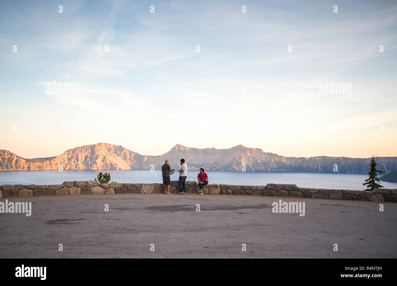 Fernsicht auf drei junge Männer sitzen auf einem kurzen Rock an der Wand mit Blick auf einen riesigen See bei Sonnenuntergang, Crater Lake, Oregon, USA Stockfoto