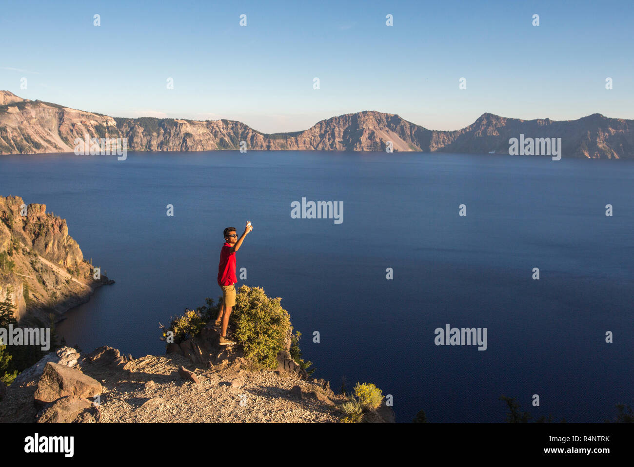 Ein junger Mann in einem roten T-Shirt stellt für eine selfie auf einem Felsvorsprung hoch über einem tiefblauen See umgeben von Bergen, Crater Lake, Oregon, USA Stockfoto