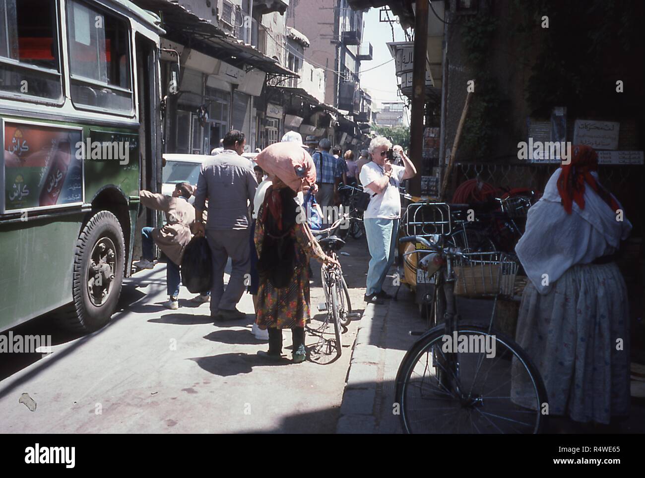 Szene einer Gasse mit Fußgänger- und Fahrzeugverkehr, in Damaskus, Syrien, Juni 1994 gefüllt. Im Rahmen einer westlichen Touristen fotografiert den Marktstand von der Seite der Straße. Am linken Rand ein kleiner Junge steigt auf einen Stadtbus mit Werbung in Arabisch für unterschiedliche Varianten von Crush Marke Soda. () Stockfoto