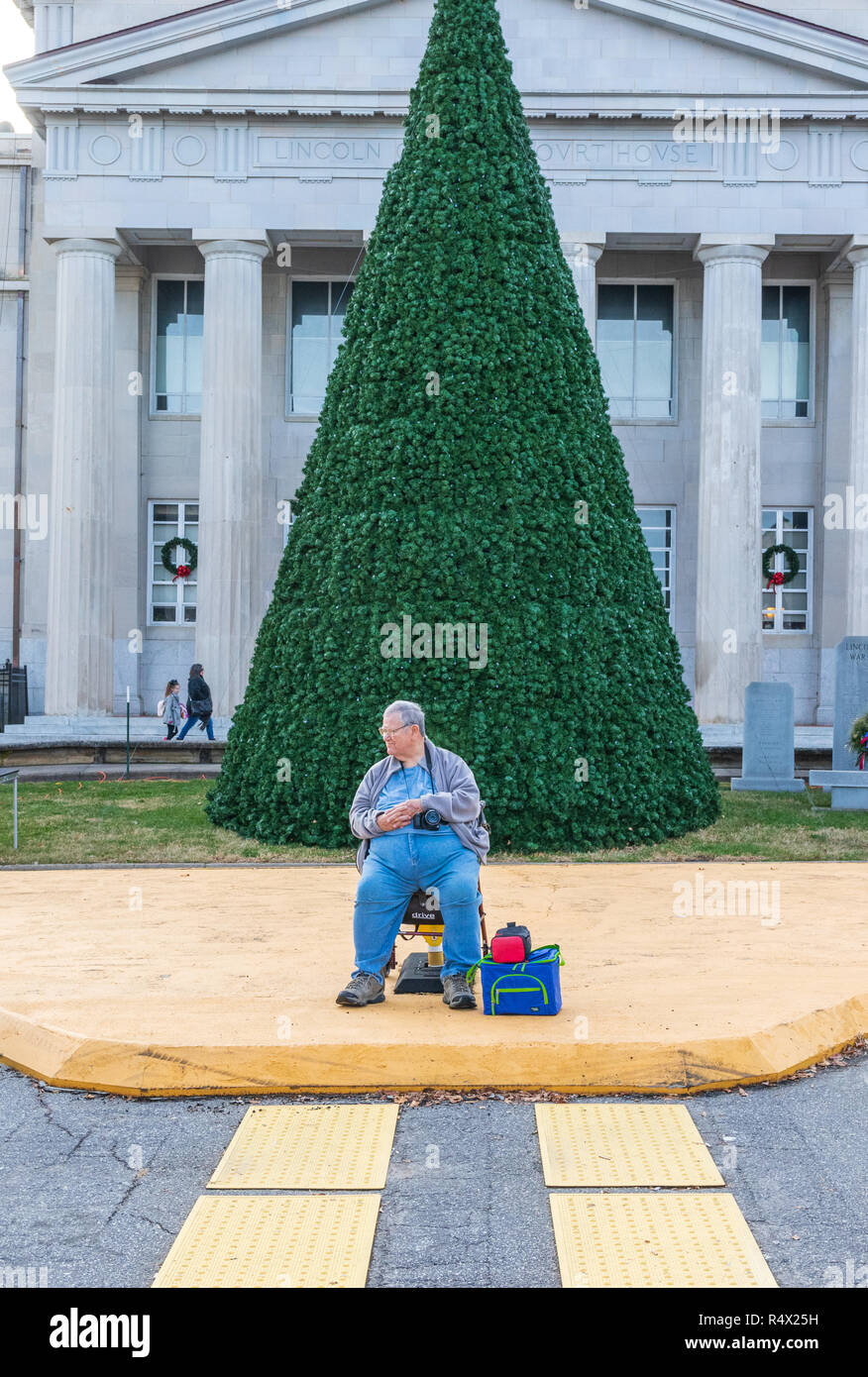 Mebane, NC, USA -11/25/18: Übergewicht Mann mit Kamera sitzt vor dem Undekorierten Weihnachtsbaum, Warten auf Parade. Stockfoto