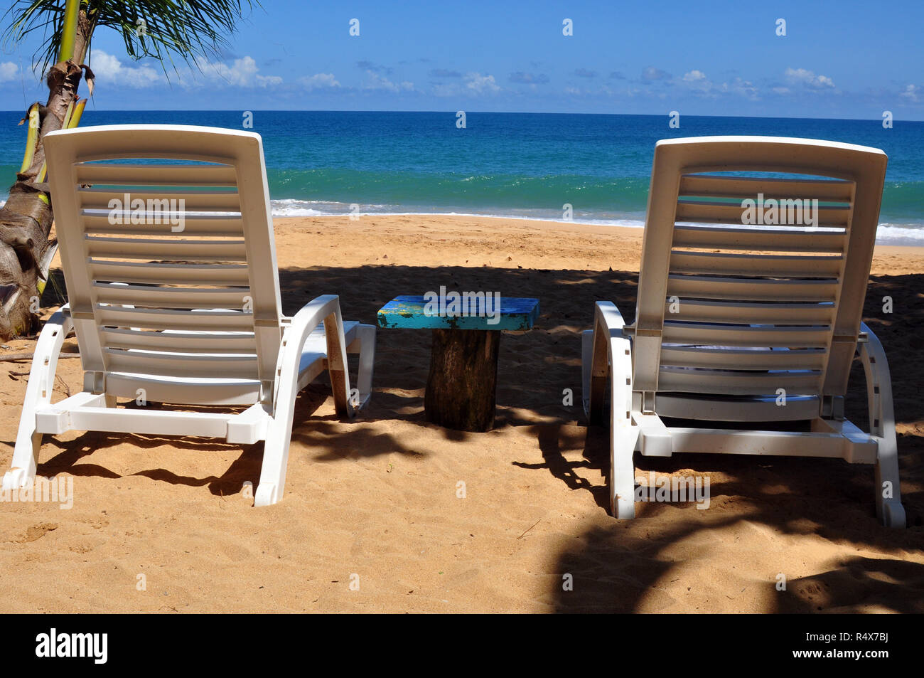 Zwei weißen Liegestühlen am Meer mit einem kleinen blauen Tisch dazwischen. Stockfoto