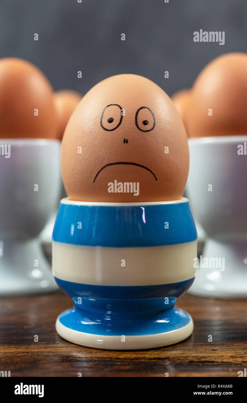 Traurig emotion Depression Konzept einer einzelnen gekochtes Ei stirnrunzelnd in verschiedenen blau gestreiften Eierbecher von Eiern in Weiß Eierbecher umgeben auf einer hölzernen t Stockfoto