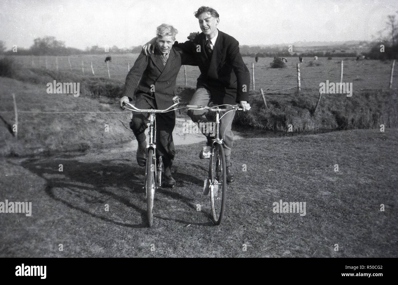 1950, historische, zwei gut gekleidete junge Männer sitzen auf ihren Fahrrädern nebeneinander auf einem grasbewachsenen Rand in der Landschaft, England UK. Stockfoto