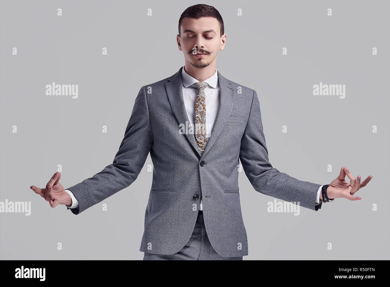 Portrait von gutaussehenden Jungen zuversichtlich arabischer Geschäftsmann mit ausgefallenen Schnurrbart in Mode grauen kompletten Anzug Meditation über studio Hintergrund Stockfoto