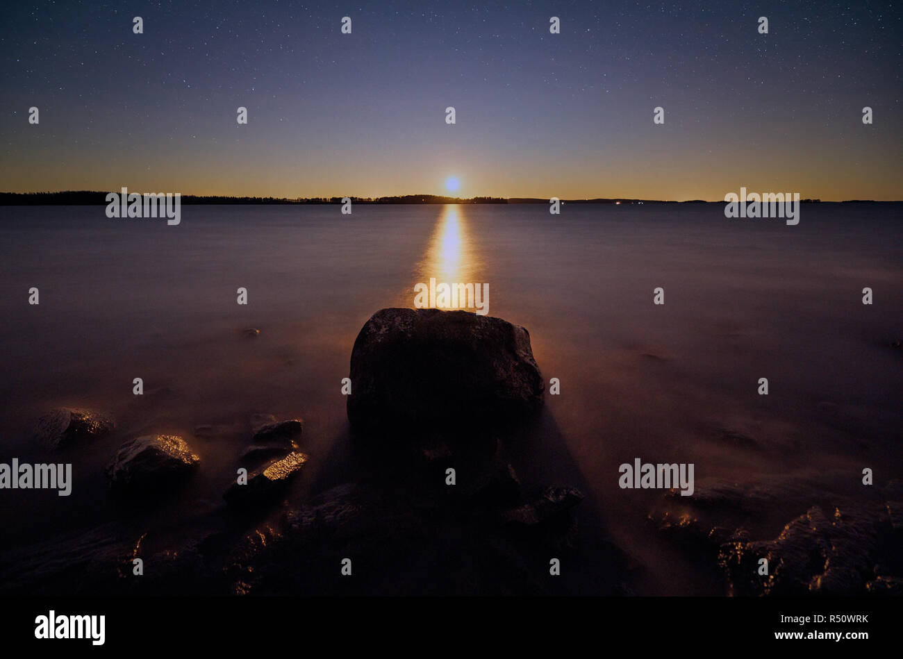 Mond scheint knapp über dem Horizont in der Nacht. Big Stone auf noch Wasser der friedlichen See in Finnland. Stockfoto