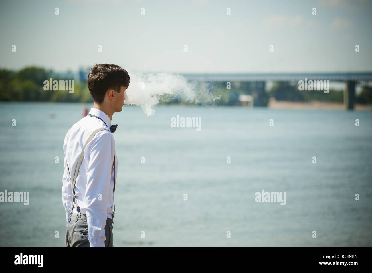 Junge dünne Mann, elegant und stilvoll in weißem Hemd, Hose mit Hosenträgern und Bow Tie. vaper Kerl raucht eine e-Zigarette, releas Rauchschwaden, gegen den Hintergrund von Fluss und Brücke Stockfoto