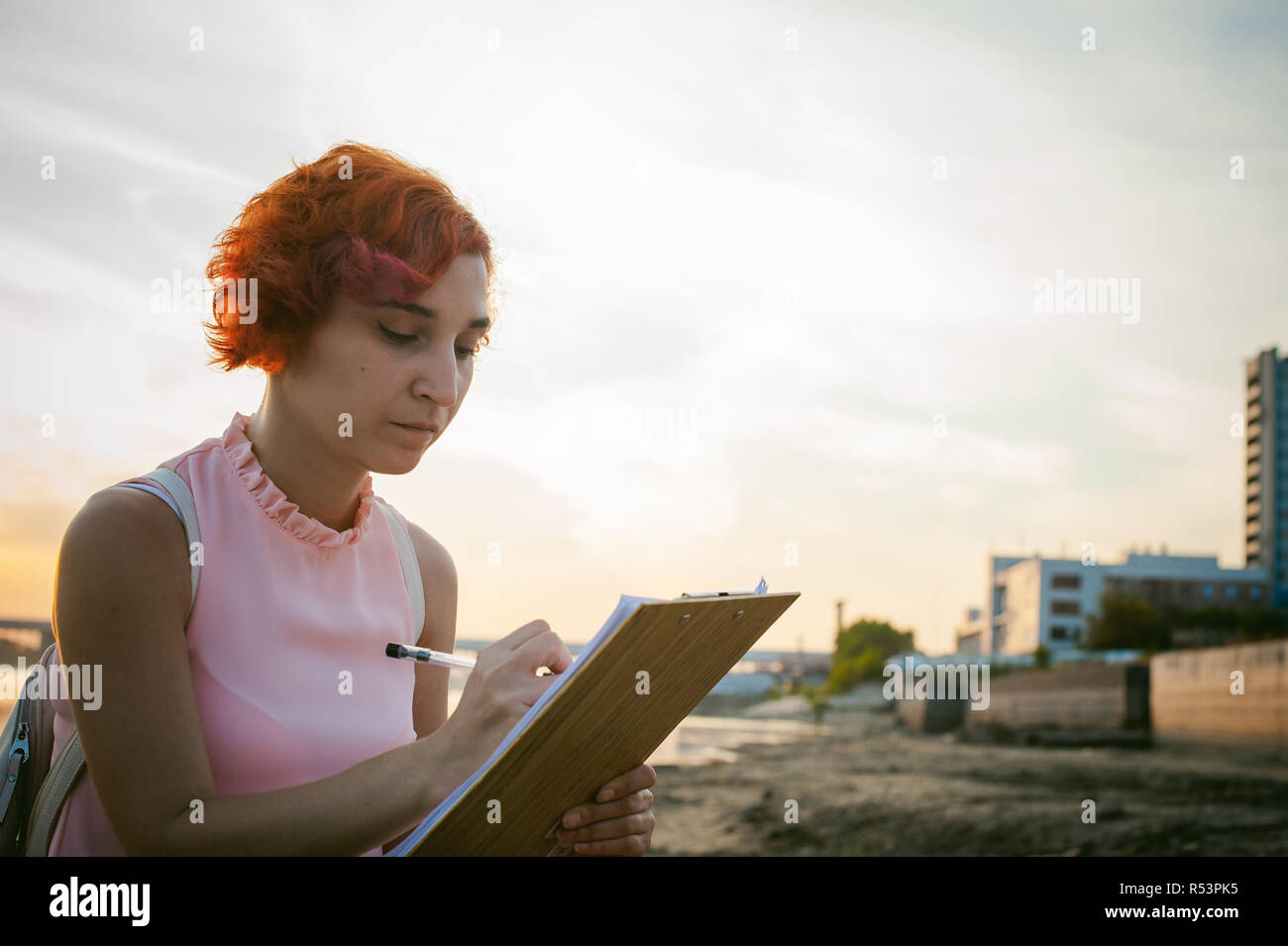 Frau mit rot gefärbten Haaren in einem blassen rosa Kleid mit weißen Rucksack, wichtige Dokumente unterzeichnet Sitzen im Freien in der Nähe des Flusses bei Sonnenuntergang, gegen einen Bootsliegeplatz Stockfoto