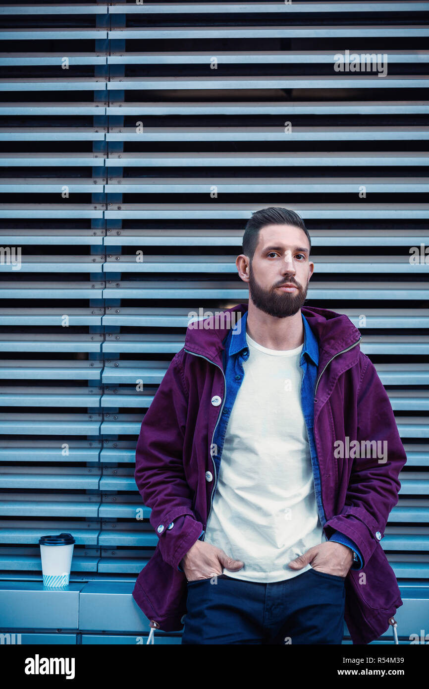 Im Herbst oder Winter Portrait von Stattlichen hipster Mann mit Bart, weisses T-Shirt, blau Shirt und kastanienbraunen Jacke Holding heiße Tasse Kaffee. Gerippte urba Stockfoto