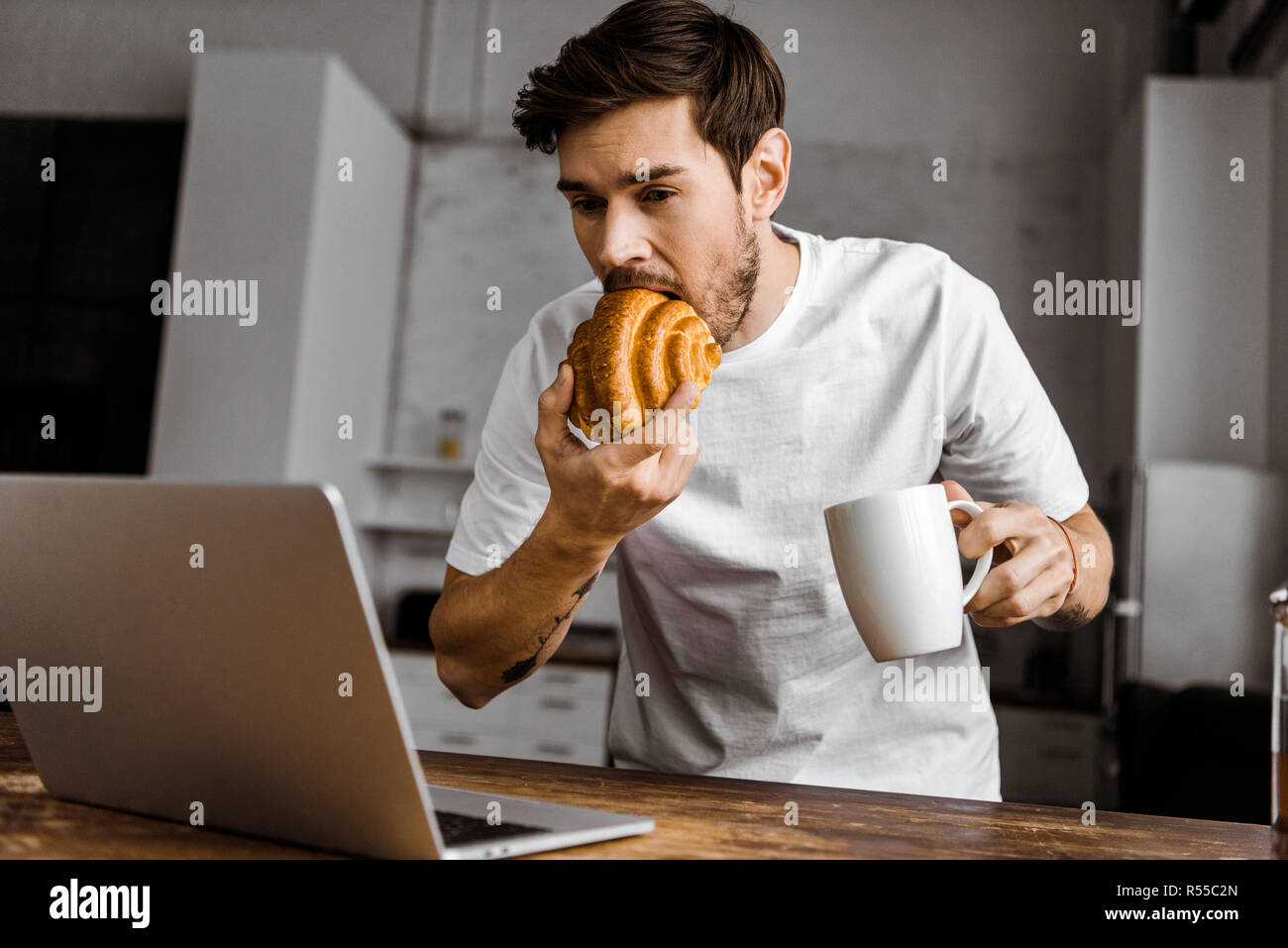 Hungrigen jungen Freelancer bei einer Tasse Tee, Croissant und Laptop arbeiten an Küche zu Hause Stockfoto