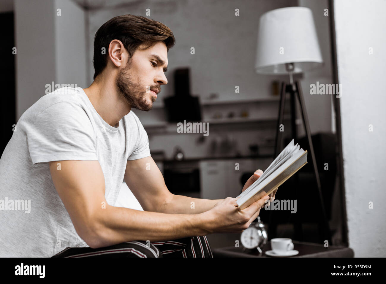 Seitenansicht des gutaussehenden jungen Mann im Schlafanzug sitzen und lesen Buch zu Hause Stockfoto