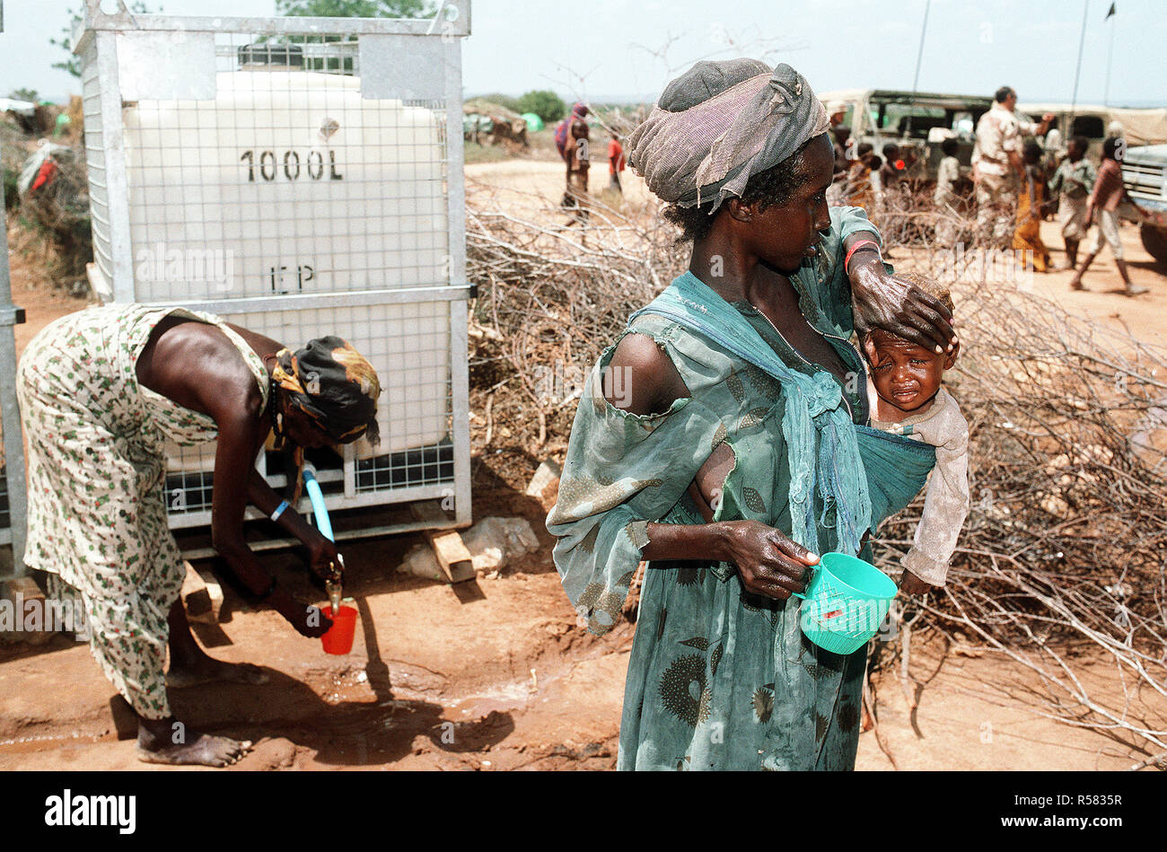 Ein Somalischer Flüchtling Frau hält Ihr Kind, wie eine andere Frau Wasser aus einem Spender im Hintergrund bekommt eine Beihilfe von bis während der Operation Restore Hope Hilfsmaßnahmen. Stockfoto
