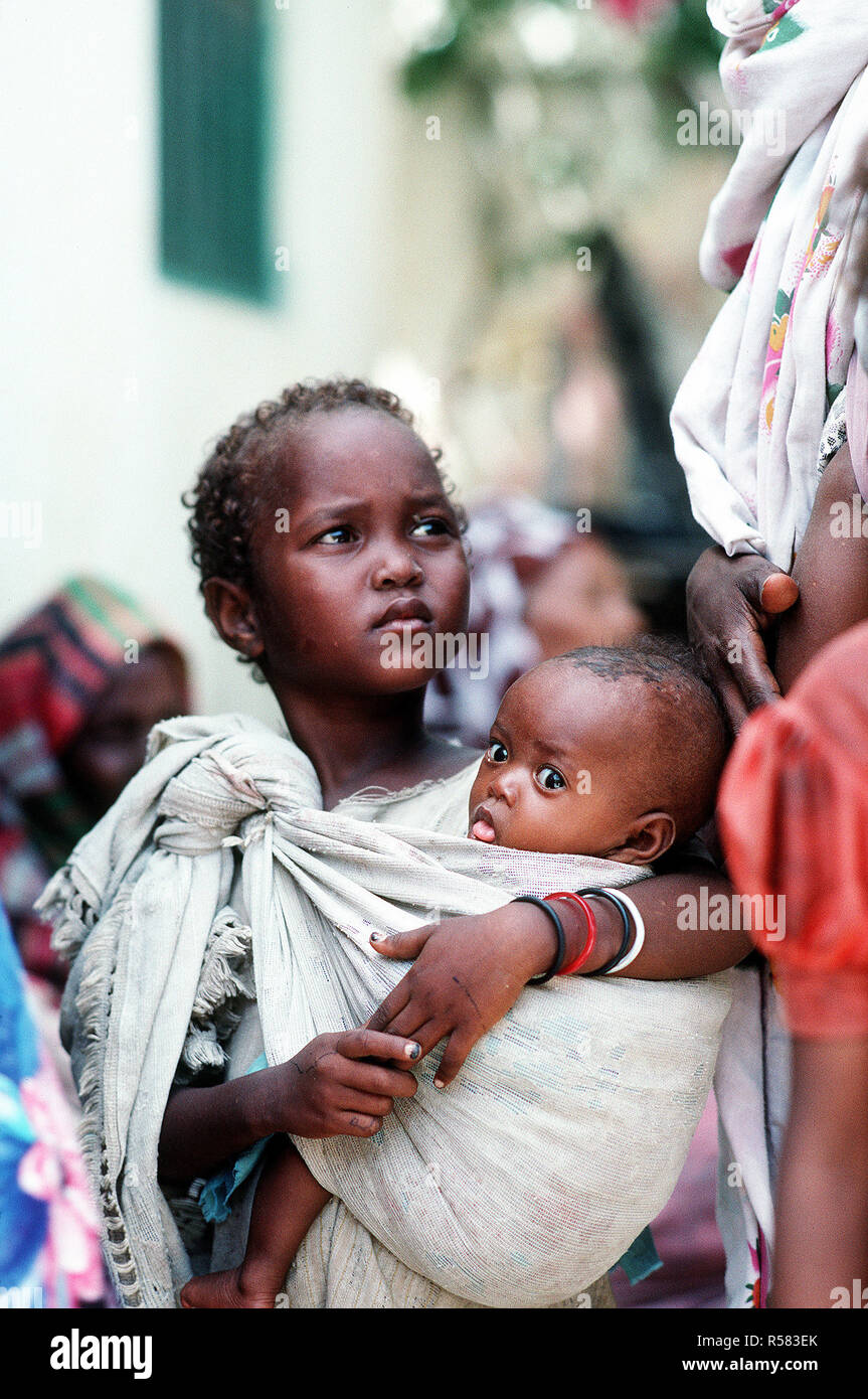 1993 - eine somalische Mädchen hält ein Kind während der Wartezeit durch ein Navy corpsman untersucht werden. Bekämpfung Service Support Abteilung 15 (CSSD-15) ist die Durchführung einer medizinischen civic Aktionsprogramm in den Straßen der Stadt während der multinationalen Entlastung Bemühung Operation Restore Hope. Stockfoto
