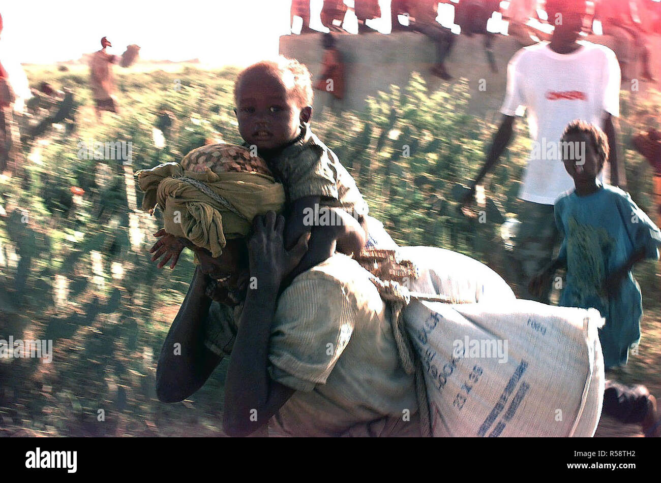 1993 - Linke Seite Profil eines somalischen Frau ihr Kind tragen und ein 50 kg Sack Mais auf ihrem Rücken als Sie geht nach rechts hinter der Kamera nach links. Unscharfe Bilder von anderen Somalis sind im Hintergrund zu sehen. Die Lebensmittel und Vorräte sind in der direkten Unterstützung der Operation Restore Hope. Stockfoto