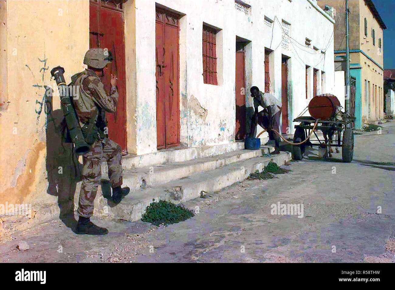 1993-A Marine steht Zurück zur Kamera, das Rauchen einer Zigarette neben einer Reihe von Gebäuden. Er hat eine M136 (4) Licht Anti-Armor Waffe umschlungen über der linken Schulter. Er wacht Ein somalischer Mann Wasser von einem Pferd liefern - Wassertank zu einem lokalen Residence. Stockfoto