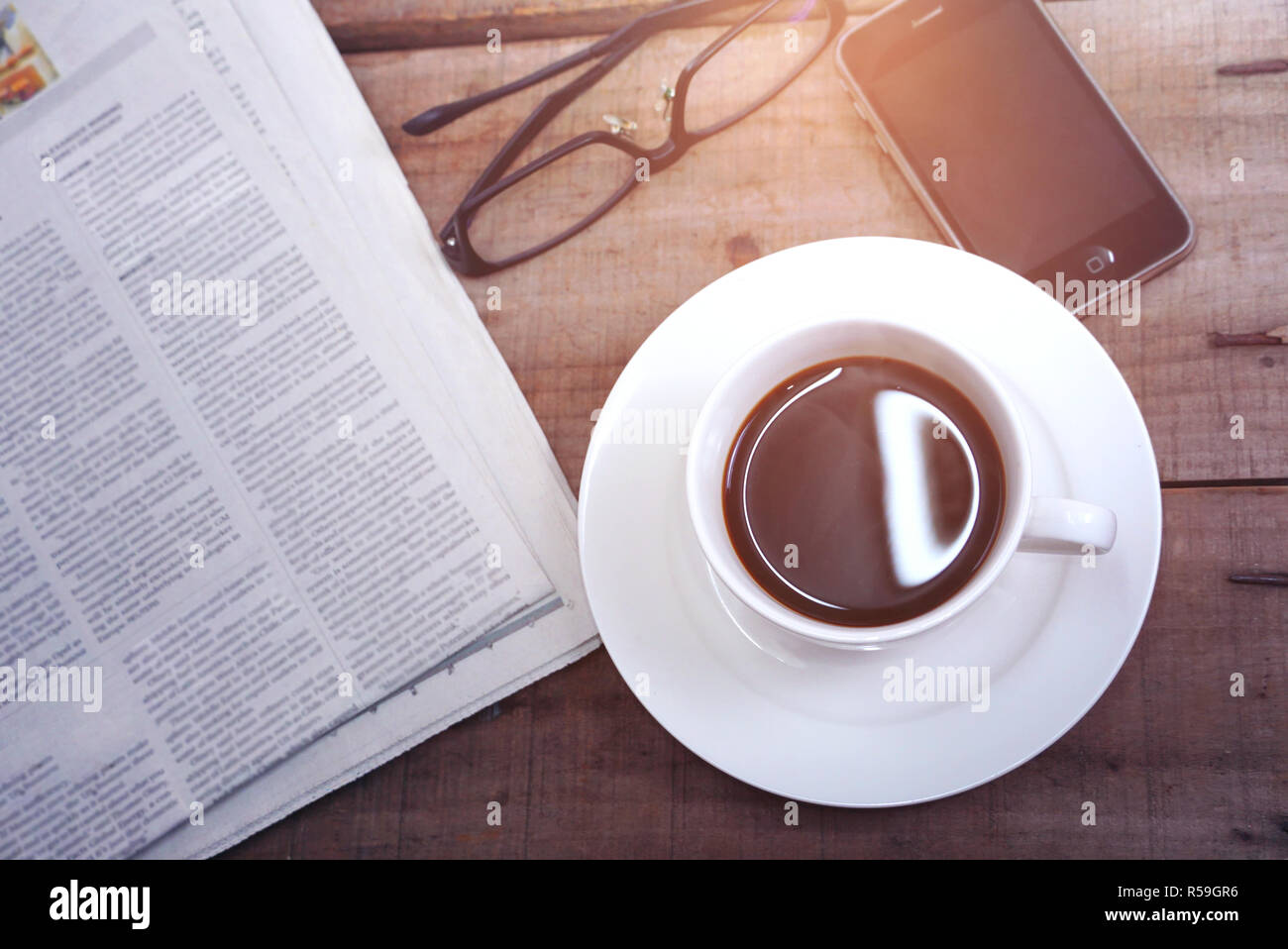 Topview Kaffeetasse mit Zeitung und Gläser und Smart Phone auf dem Tisch mit Sonnenlicht aus dem Fenster Stockfoto
