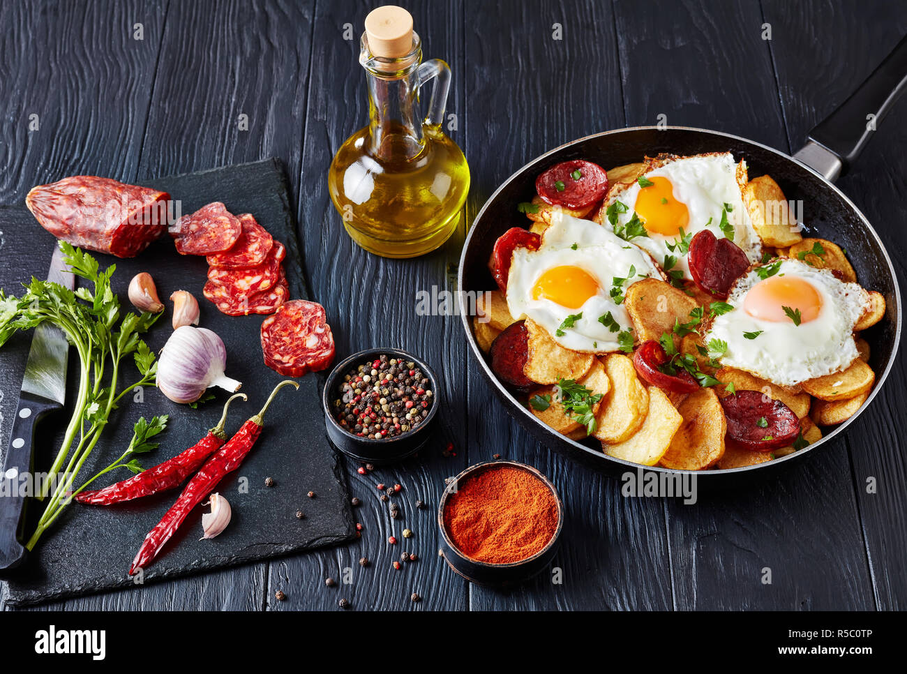 Traditionelle spanische Huevos rotos oder Huevos estrellados - gebratene Eier mit Kartoffeln, Würstchen Chorizo in einer Pfanne auf einem schwarzen Holztisch mit Ingr Stockfoto