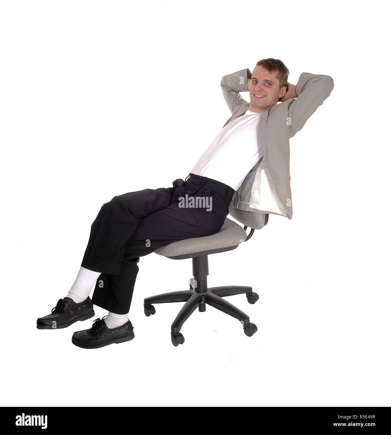 Ein junger Mann in dunklen Hosen und eine graue Jacke sitzen auf einem Bürostuhl mit seine Hand hinter seinem Kopf, auf weißem Hintergrund Stockfoto
