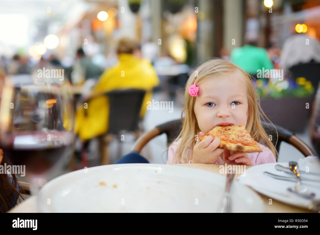 Nette lustige kleine Mädchen essen ein Stück Pizza im Restaurant im Freien im Sommer am Abend. Kinder essen Junk Food. Stockfoto