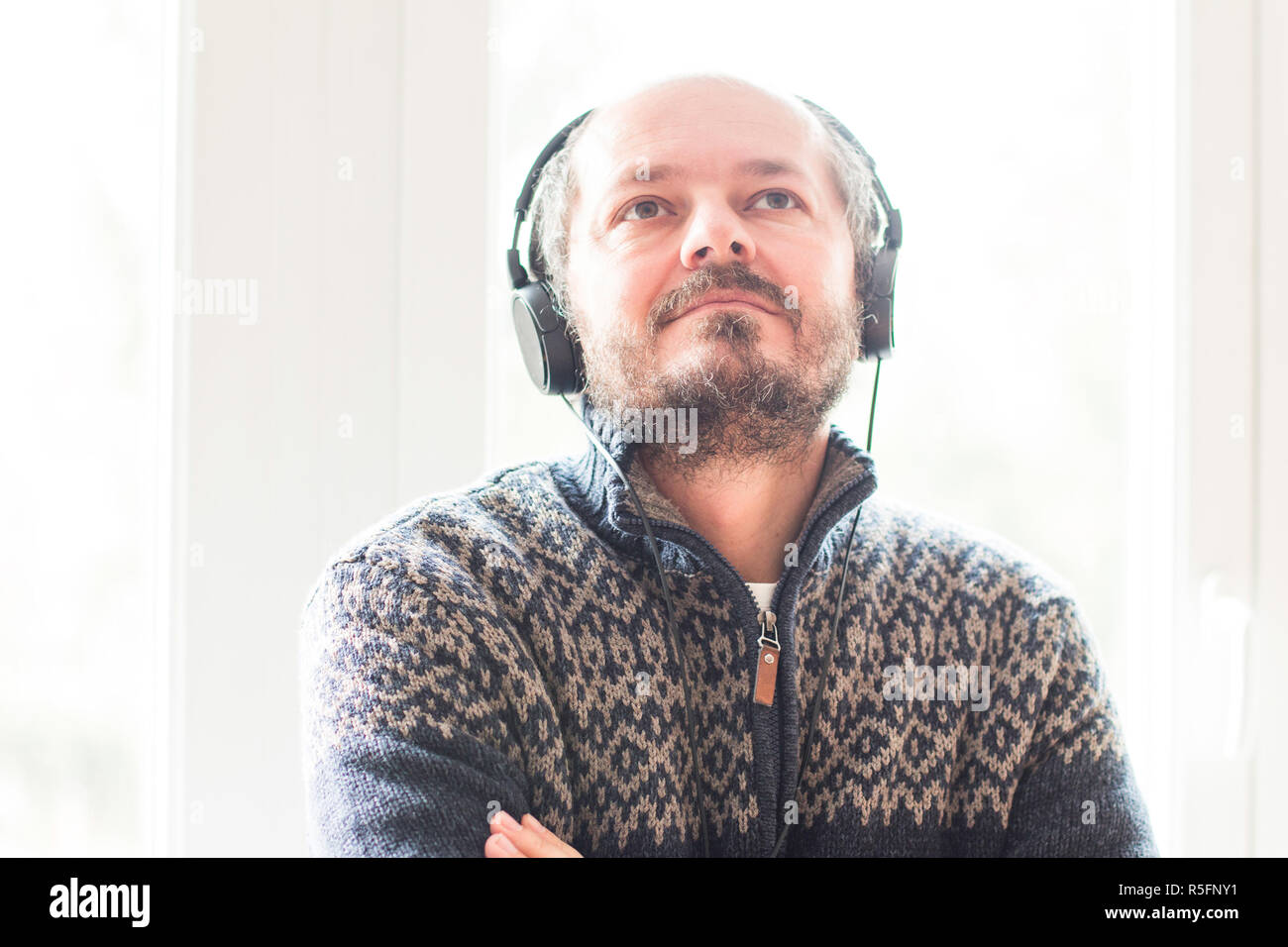 Lächelnd mittleren Alter Mann Musik hören mit Kopfhörern beim Stehen am Fenster zu Hause, Kopf und Schultern, Musik Konzept Stockfoto