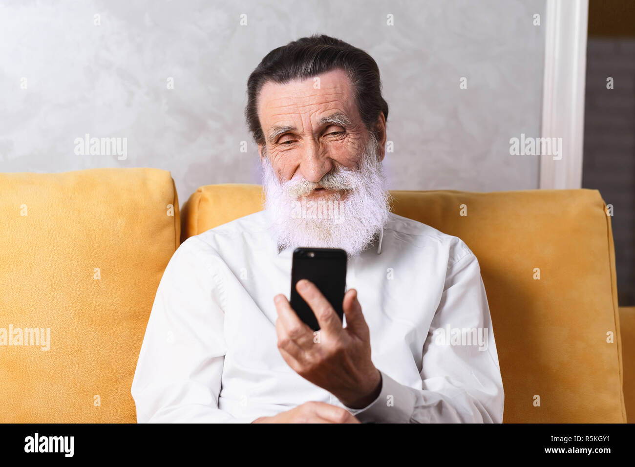 Gerne älteren bärtigen Mann mit weißem T-Shirt sitzen auf dem gelben Sofa und sie ihr Smartphone Stockfoto