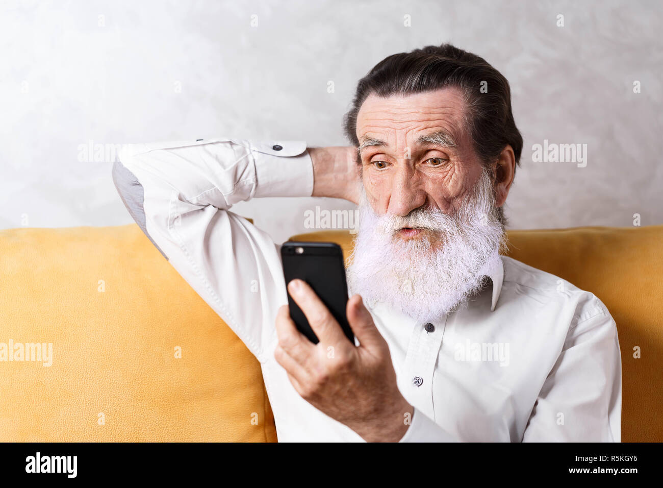 Überrascht älterer bärtiger Mann mit weißem T-Shirt sitzen auf dem gelben Sofa und Suchen auf dem Bildschirm seines Smartphones Stockfoto