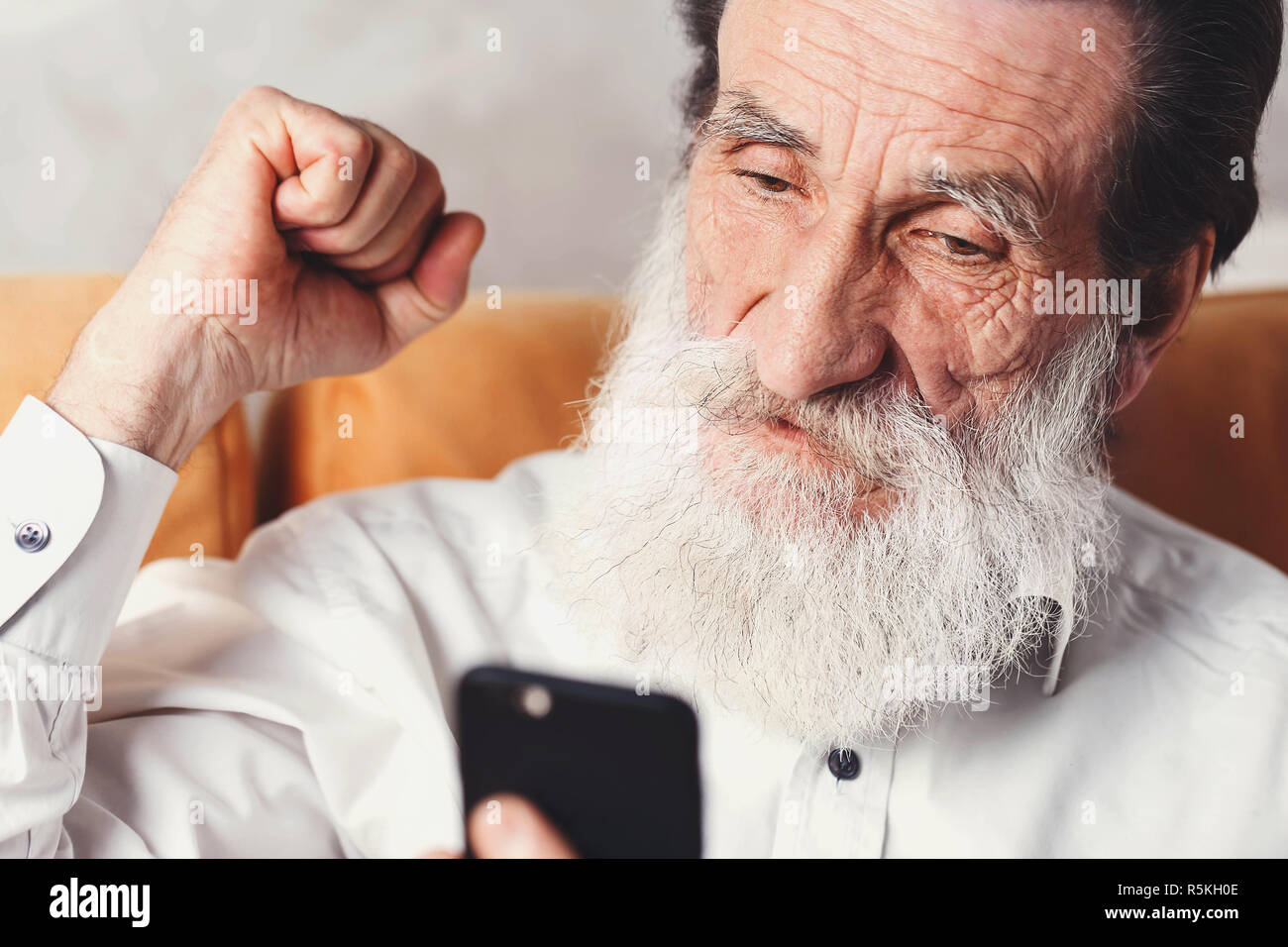 Moderne älterer bärtiger Mann mit weißem T-Shirt sitzen auf dem gelben Sofa und Jubel von etwas auf die sein Smartphone Stockfoto