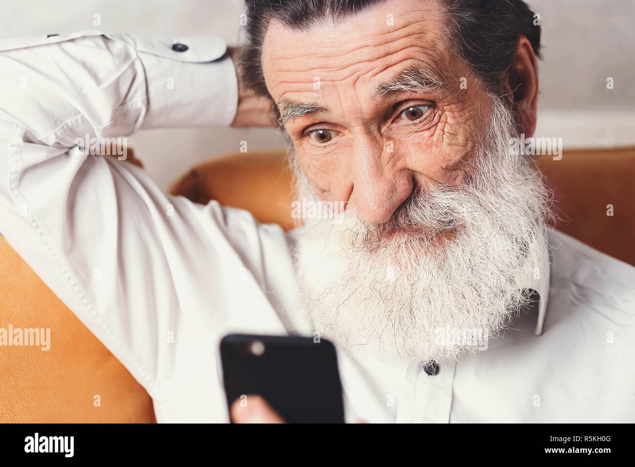 Moderne älterer bärtiger Mann mit weißem T-Shirt sitzen auf dem gelben Sofa und sie ihr Smartphone, die er am Telefon mit der Erwartung, den er benötigt News Stockfoto