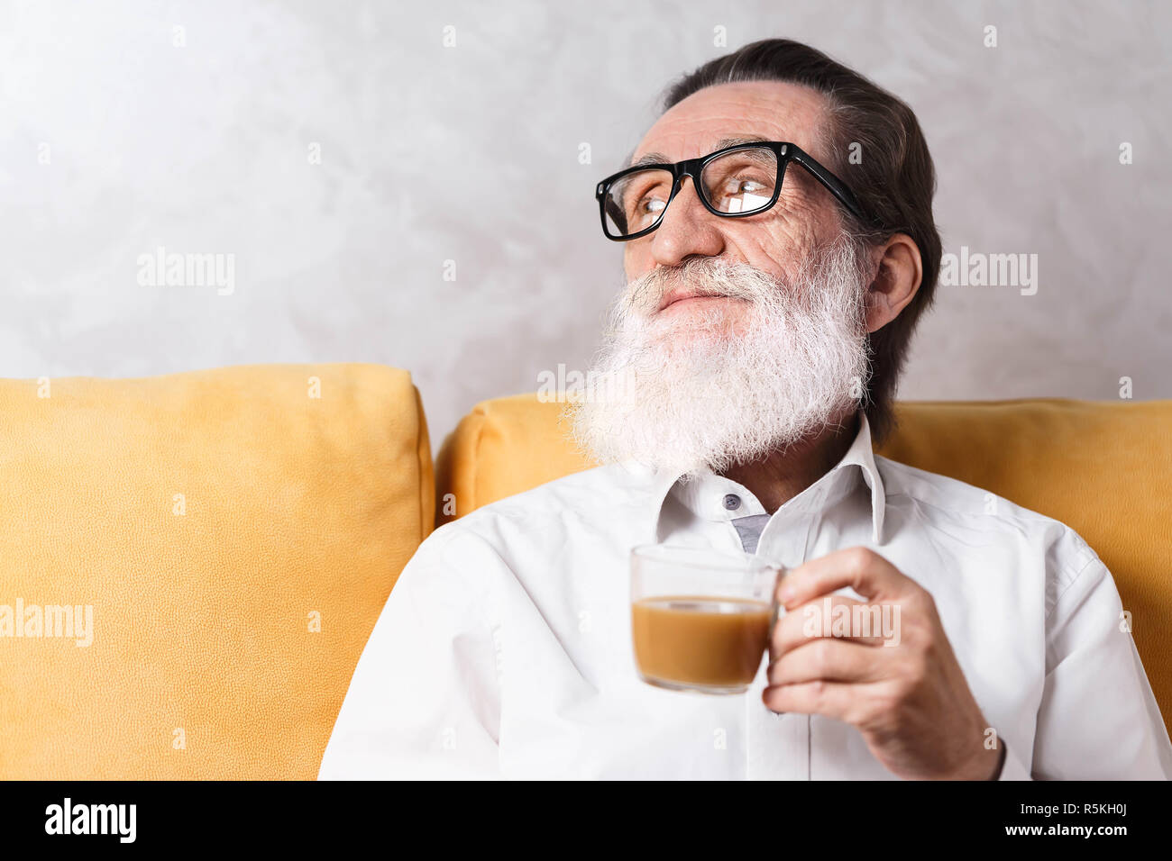 Charmante älterer bärtiger Mann im weißen Hemd mit Brille sitzen auf dem gelben Sofa und trinkt seinen Kaffee am Morgen Stockfoto