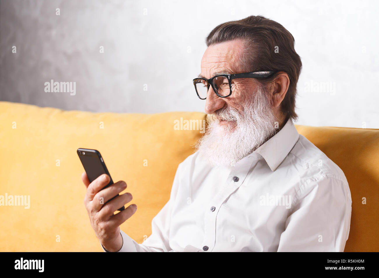 Charmante älterer bärtiger Mann im weißen Hemd mit Brille sitzen auf dem gelben Sofa und Lesen von Nachrichten auf dem Smartphone Stockfoto