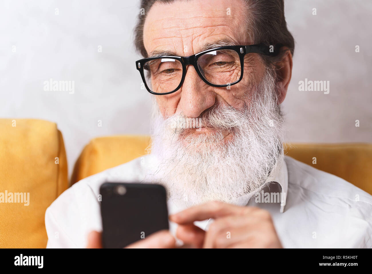 Charmante älterer bärtiger Mann im weißen Hemd mit Brille sitzen auf dem gelben Sofa und scrollen Facebook Stockfoto