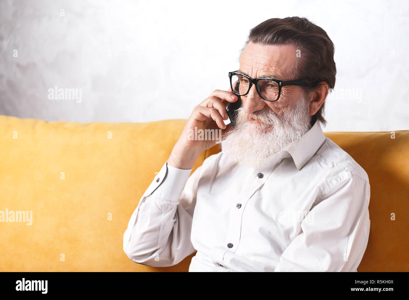 Schöne bärtige älterer Mann im weißen Hemd mit Brille plaudern auf ein Mobiltelefon beim Entspannen auf einer gelben Sofa in seinem Lichte Wohnzimmer Stockfoto