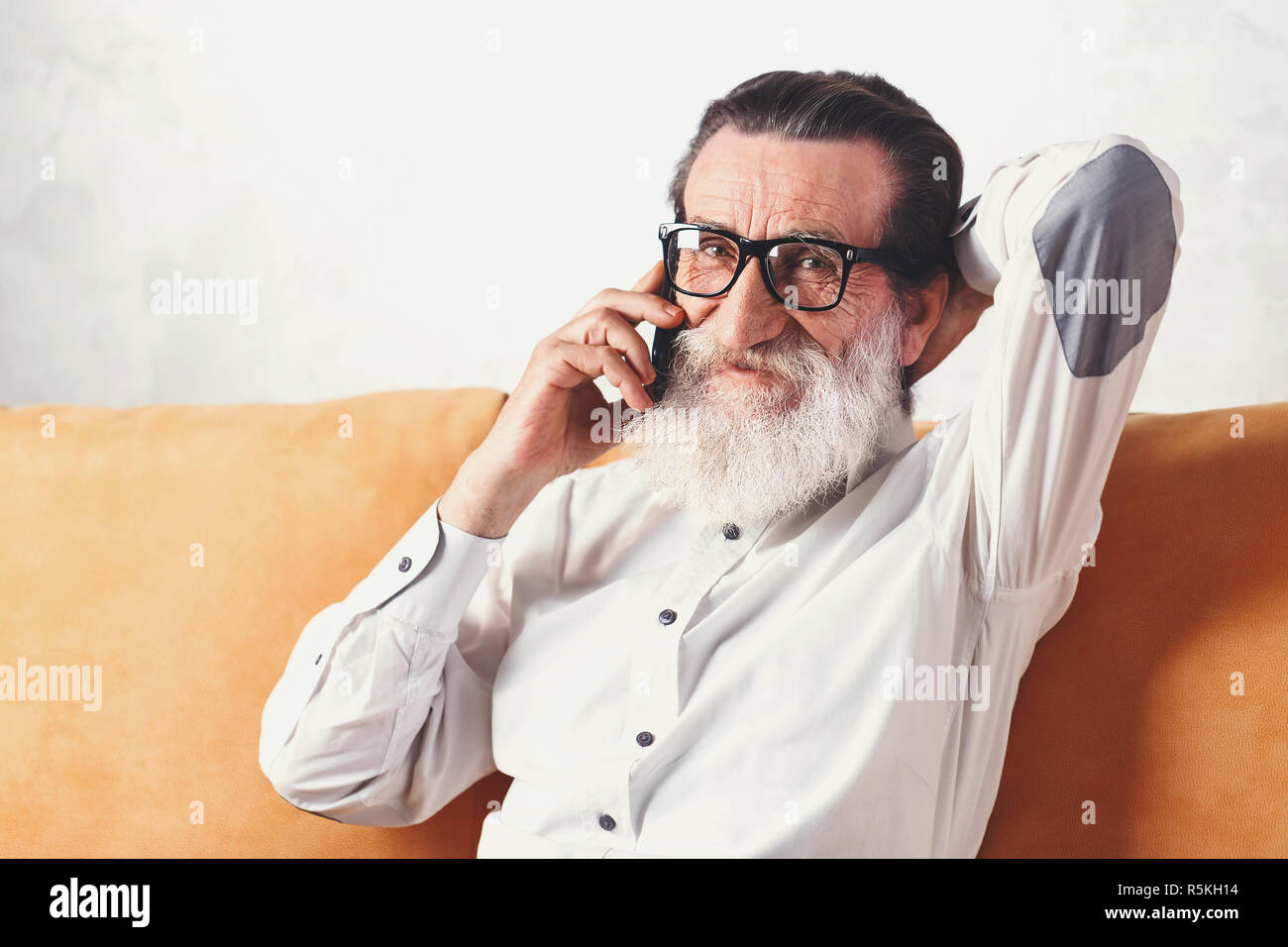 Portrait von gut aussehenden Bärtigen älteren Mann im weißen Hemd mit Brille plaudern auf ein Mobiltelefon beim Entspannen auf einer gelben Sofa in seinem Lichte Wohnzimmer, er schaut in die Kamera Stockfoto