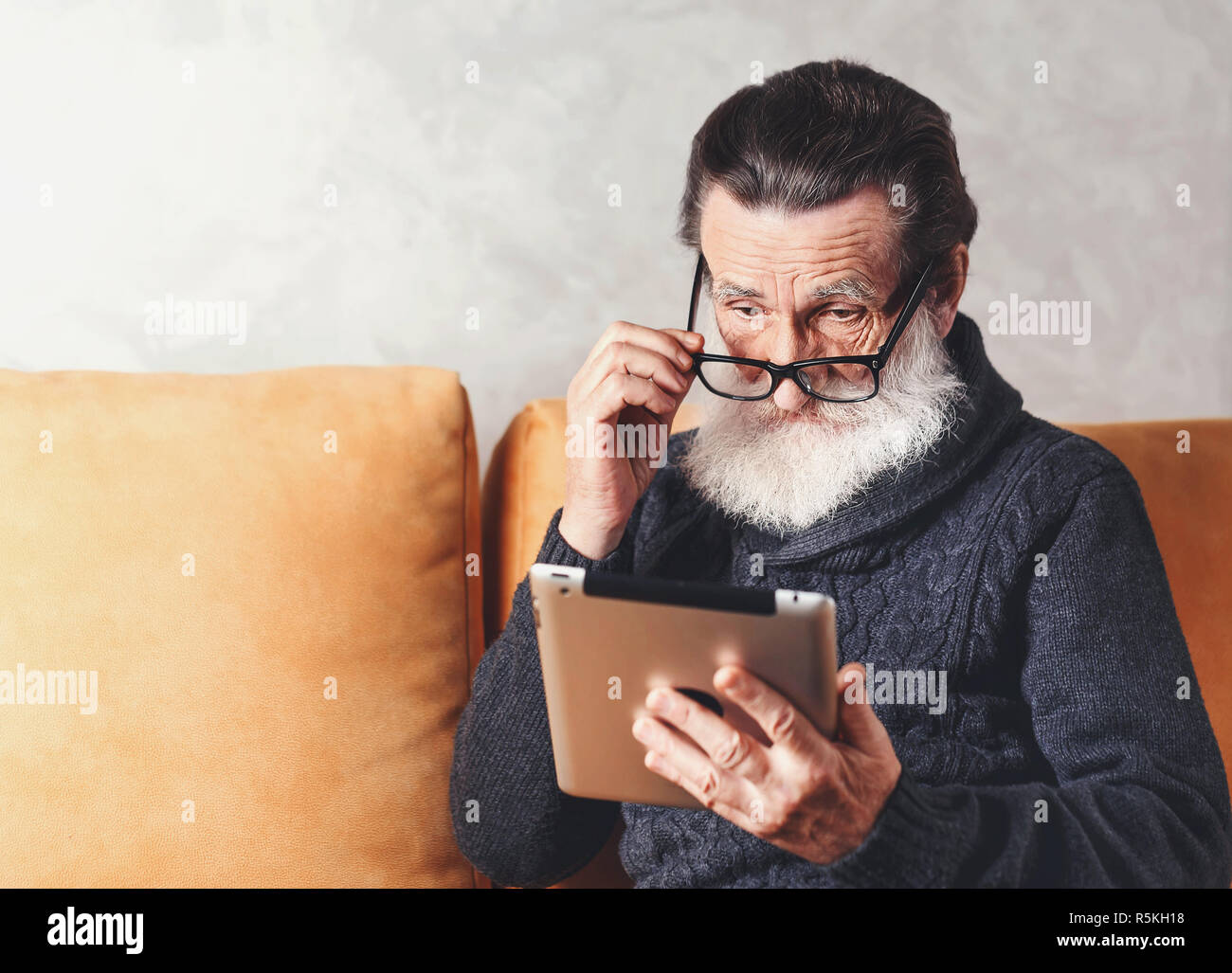 Clevere älterer bärtiger Mann mit Brille grau Pullover lesen elektronisches Buch auf dem digitalen Tablet beim Sitzen auf einem gelben Sofa in seinem Lichte Wohnzimmer, er über seine Brille Stockfoto
