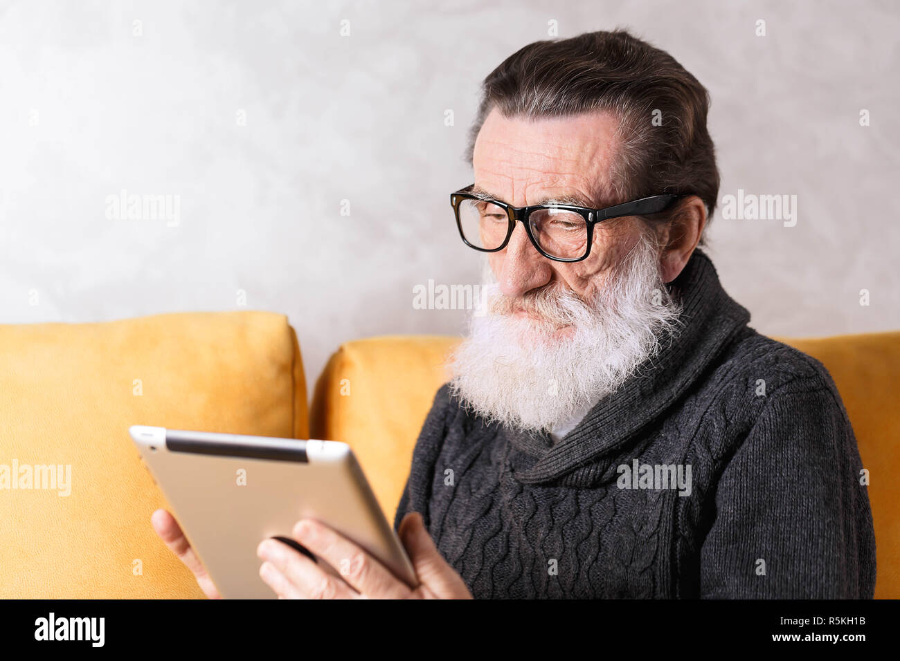 Clevere älterer bärtiger Mann mit Brille grau Pullover lesen elektronisches Buch auf dem digitalen Tablet beim Sitzen auf einem gelben Sofa in seinem Lichte Wohnzimmer, konzentrierte er sich auf die Lesung Stockfoto