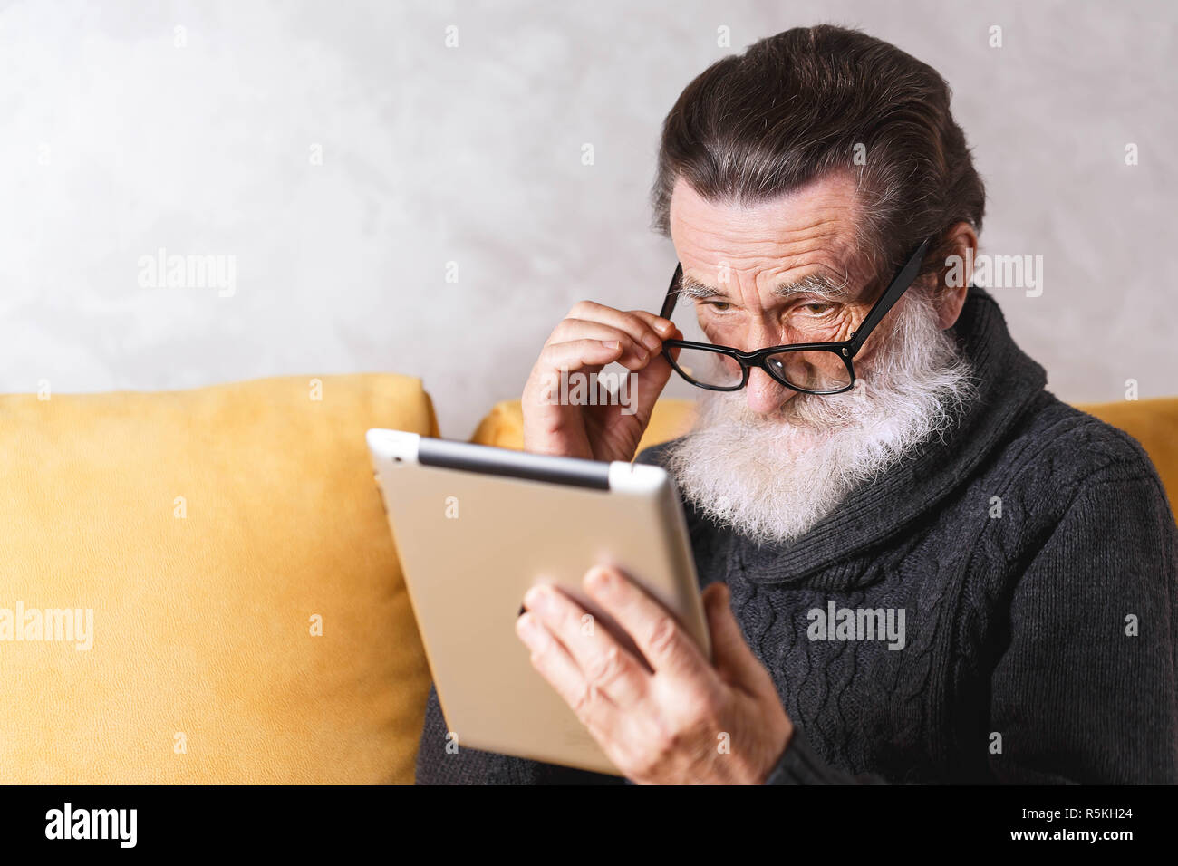 In älteren bärtigen Mann mit Brille grau Pullover sitzen auf einem gelben Sofa in seinem Lichte Wohnzimmer und Blick auf seine Brille und etwas lesen auf dem Tablet-PC. Stockfoto