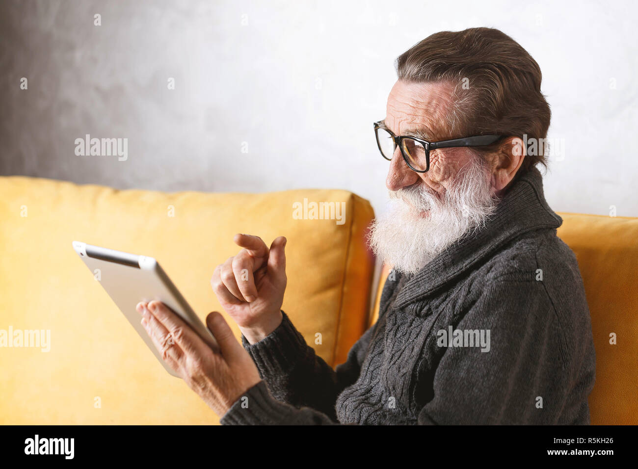 In älteren bärtigen Mann mit Brille grau Pullover sitzen auf einem gelben Sofa in seinem Lichte Wohnzimmer und der schlüsselkarte Facebook News auf der digitalen Tablet. Stockfoto