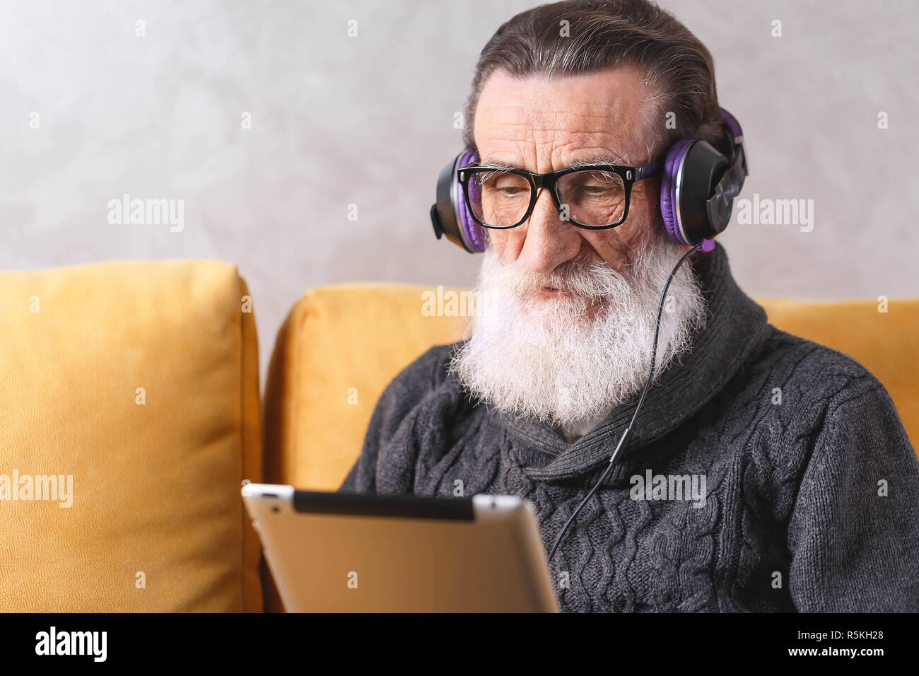 Senior zeitgenössische bärtigen Mann mit Brille grau Pullover sitzen in der Kopfhörer auf ein gelbes Sofa in seinem Lichte Wohnzimmer und interessanten Film auf dem Tablet-PC. Stockfoto