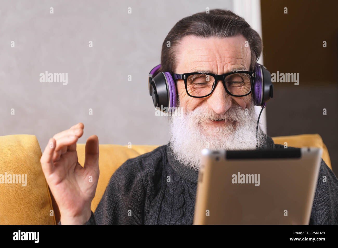 Senior heiter bärtigen Mann mit Brille grau Pullover sitzen in der Kopfhörer auf ein gelbes Sofa in seinem Lichte Wohnzimmer und Chatten mit seiner Familie auf Skype auf dem digitalen Tablet, Technik, People, Lifestyle- und Kommunikationskonzept Stockfoto