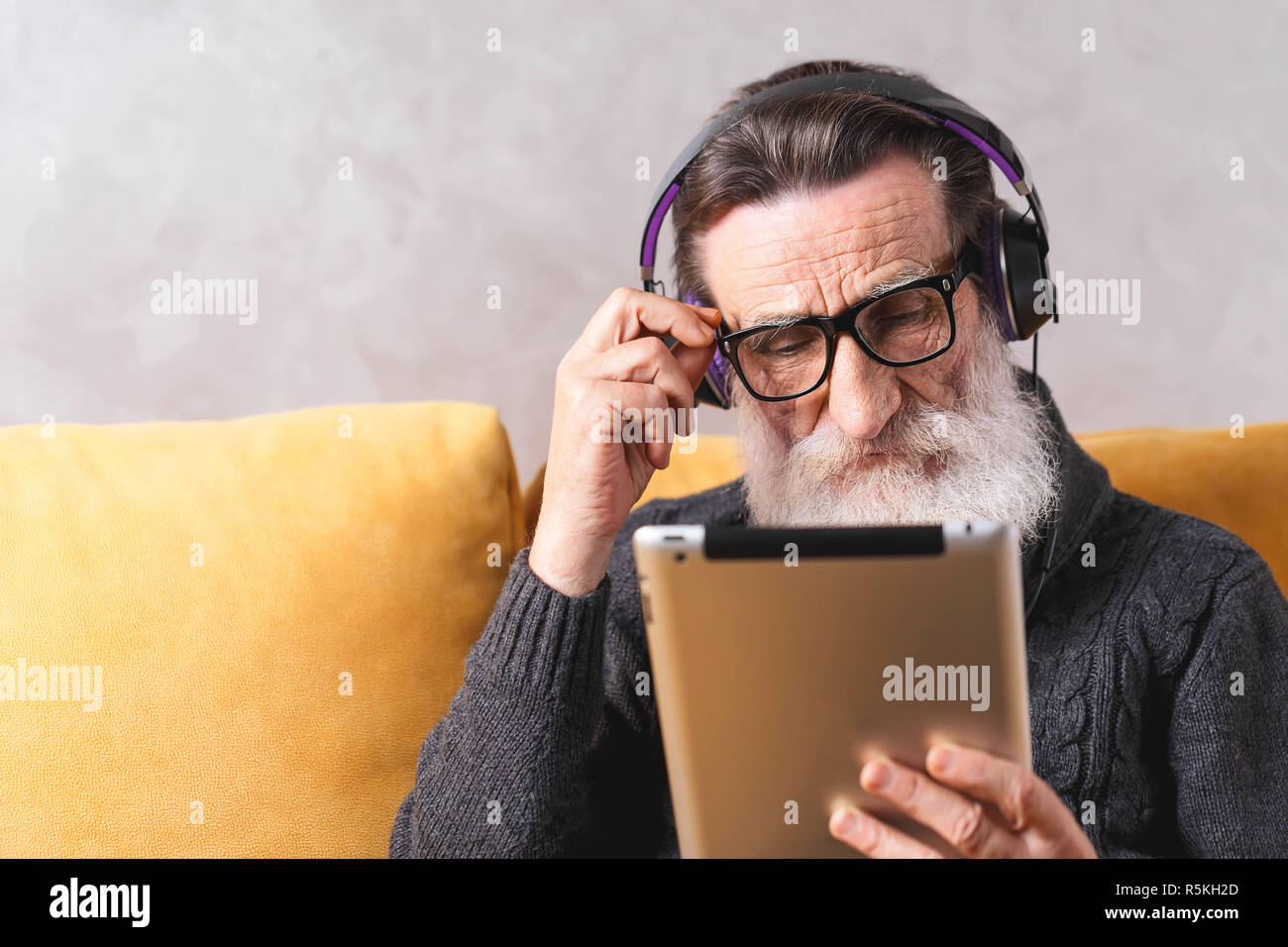 Senior Ernst bärtigen Mann mit Brille grau Pullover angespannt Angesichts der digitalen Tablet während der Sitzung in der Kopfhörer auf ein gelbes Sofa in seinem Lichte Wohnzimmer. Stockfoto