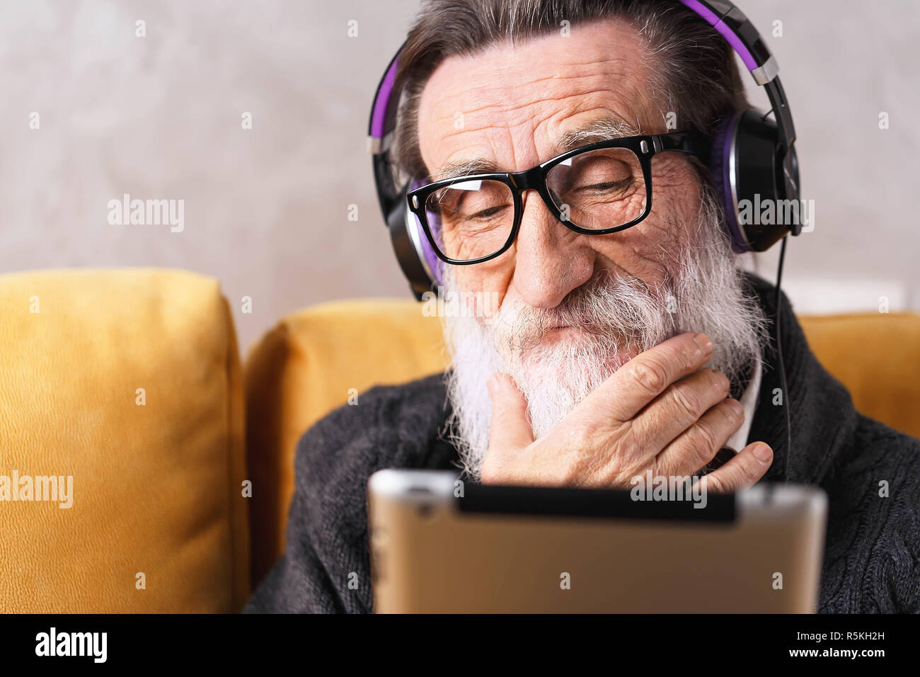 Senior nachdenklich bärtigen Mann mit Brille grau pullover Lernen der digitalen Tablet während der Sitzung in der Kopfhörer auf ein gelbes Sofa in seinem Lichte Wohnzimmer setzen, er richtet seinen Bart Stockfoto